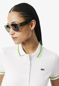 Occhiali da sole con montatura trasparente, lenti scure e aste verdi. Il modello indossa una polo bianca con accenti a righe verdi e gialle.