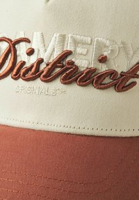 Kermainen ja ruosteenvärinen lippalakki, jossa on brodeerattu "Amery District" -teksti. Sen muoto on jämäkkä ja kangas on teksturoitu. Yksinkertaisia ompeluyksityiskohtia.