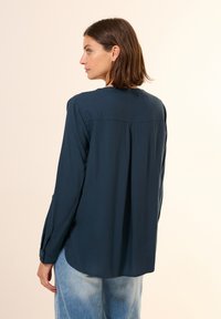 Marineblauwe longsleeve met ronde hals, zachte textuur en een plooidetail op de rug. Draag het met lichtblauwe spijkerbroek, minimaal gestyled.