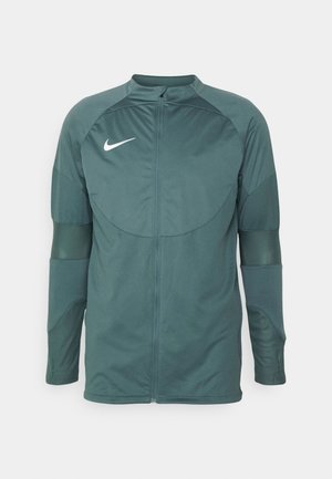 Giacca Nike di colore verde scuro, con colletto alto, zip frontale completa e pannelli testurizzati sulle maniche. Logo Nike bianco sul petto.