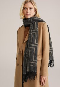 Pelēka un bēša ar rakstu šalle ar frēziem malās, izgatavota no mīksta materiāla, sakrauta uz kamieļu krāsas mēteļa, ar ģeometrisku dizainu.