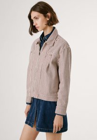 Giacca a righe in tessuto rosso e bianco, con chiusura a zip, due tasche e colletto. Abbinata a una gonna di jeans, che mostra un look casual sovrapposto.