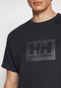 Homme portant un T-shirt noir Helly Hansen avec un logo imprimé en noir plus foncé sur la poitrine, recadré aux épaules et à la bouche.