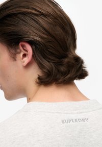 Jasnoszara bluza z teksturowanej tkaniny, okrągłym dekoltem oraz haftowanym napisem "SUPERDRY" na plecach, blisko kołnierza.