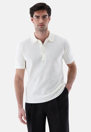 Mann trägt weißes kurzärmeliges Strick-Poloshirt mit perforierter Textur und schwarze Hose, steht mit einer Hand in der Tasche.