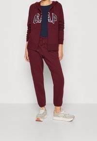 Tuta bordeaux composta da una felpa con zip e logo "GAP", abbinata a pantaloni jogger a gamba affusolata. Indossata con sneakers sportive in tonalità neutra.