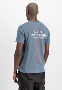 Camiseta gris de mangas cortas, con texto blanco y rojo en la parte posterior: "ALPHA INDUSTRIES", "KNOXVILLE TENNESSEE" y "REBEL BEFORE FLIGHT."