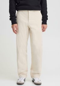 Bēša kokvilnas bikses ar brīvu piegriezumu, priekšpusē kabatas un smalka tekstūra, kopā ar melnām un baltām sporta apaviem.