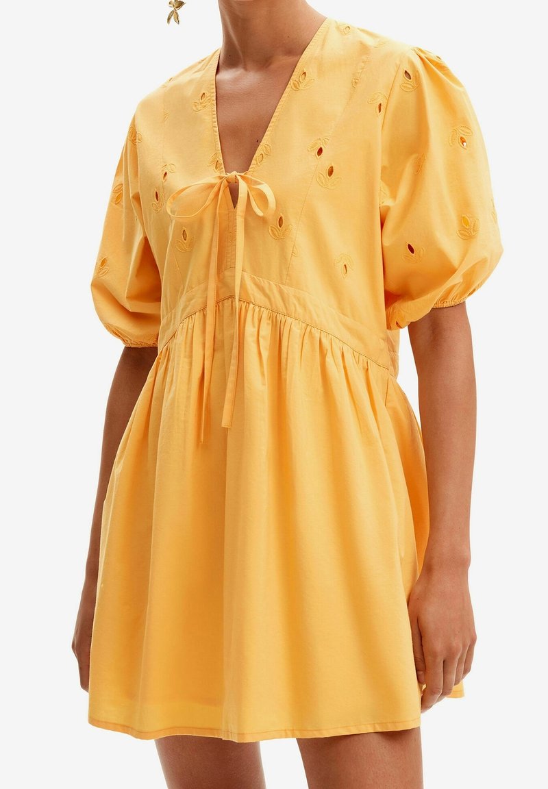 Femme portant une robe jaune ample avec des manches bouffantes, des découpes florales brodées et un lien à l'avant au niveau de l'encolure en V.