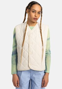 Gilet en crème matelassé avec un col en V, doté de boutons-pression et d'un passepoil contrastant. Porté par-dessus une chemise à manches longues texturée et colorée.