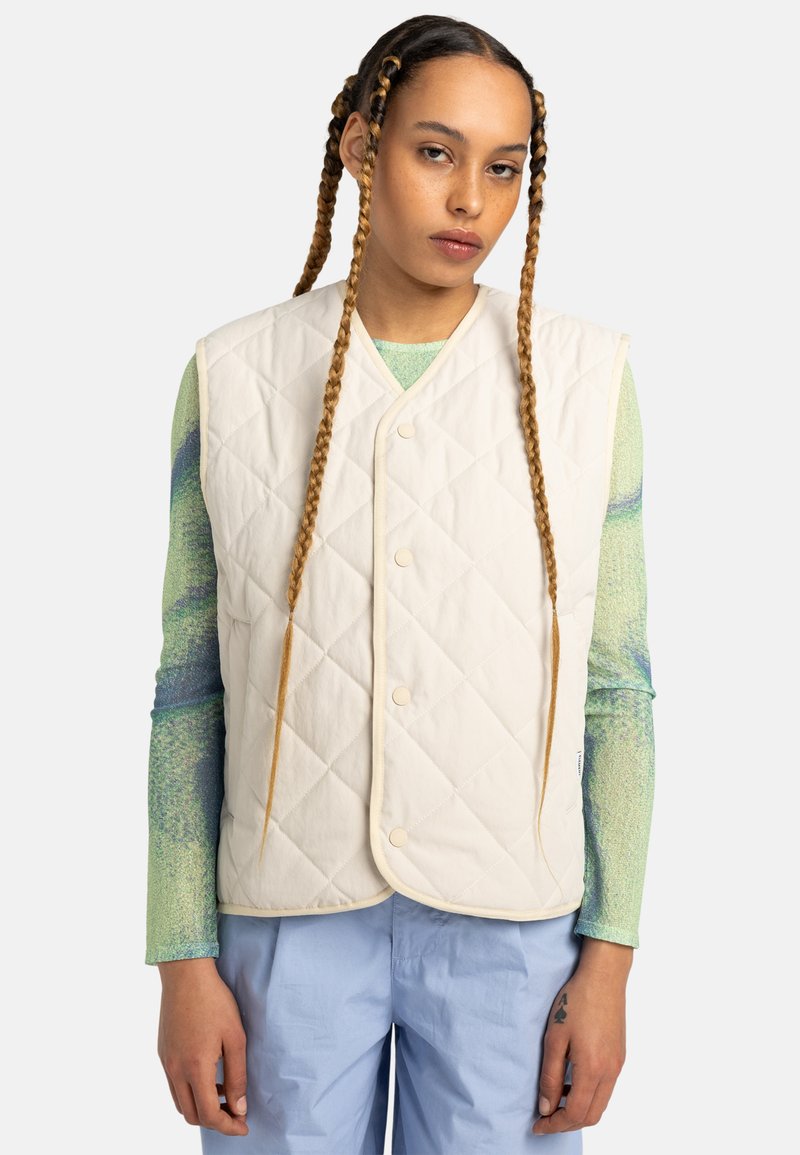 Gilet en crème matelassé avec un col en V, doté de boutons-pression et d'un passepoil contrastant. Porté par-dessus une chemise à manches longues texturée et colorée.