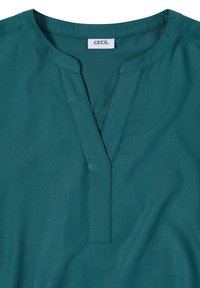 Blouse bleu sarcelle avec un col en V fendu et une étiquette blanche visible indiquant « CECIL » au niveau du col arrière.