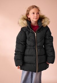 Černá péřová bunda s kapucí lemovanou umělým kožíškem, strukturovaným šitím, zapínáním na zip a staženým pasem, nošená přes skořicový top a světlé džínové kalhoty.