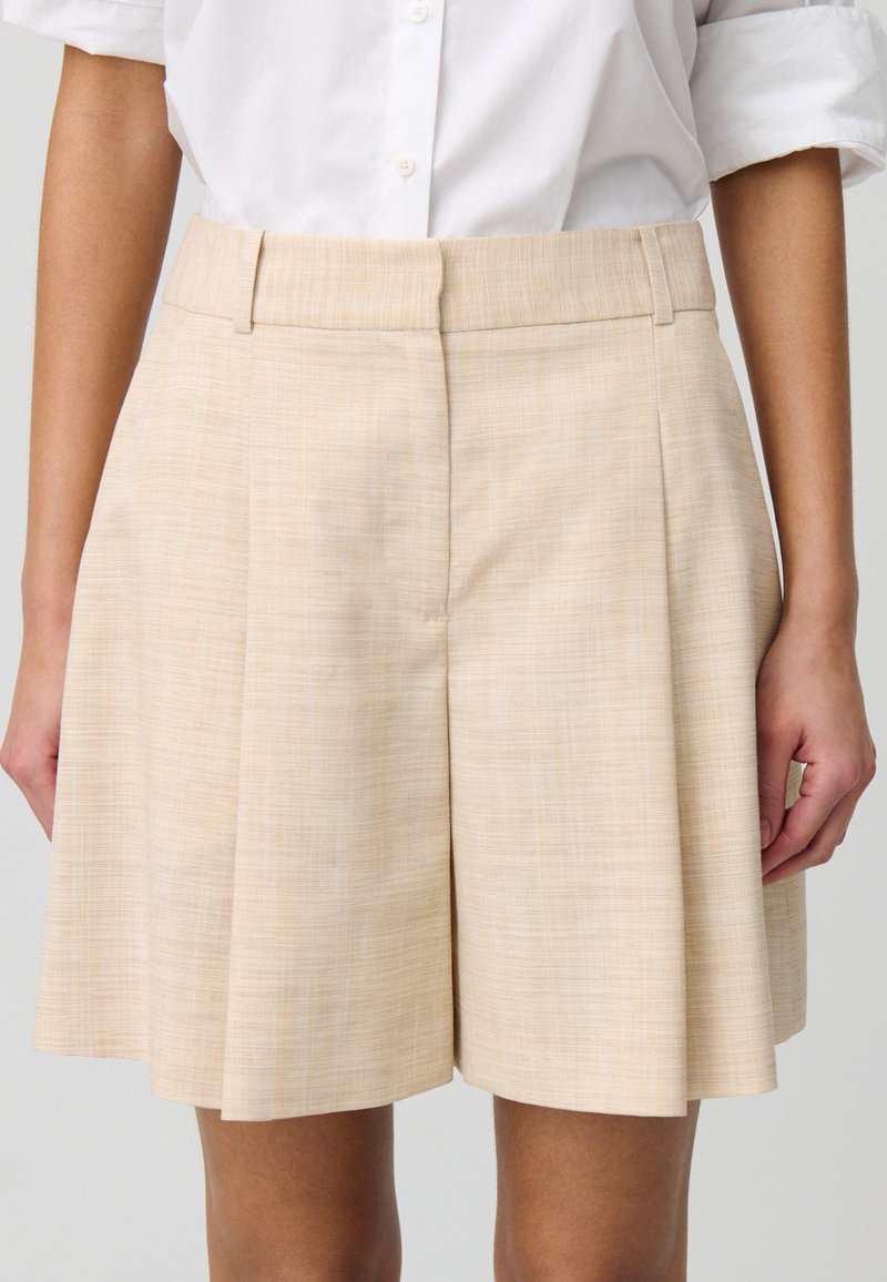 Pantaloni corti beige plissettati realizzati in un tessuto texture, con una vita piatta e tasche laterali, dal taglio ampio e morbido.