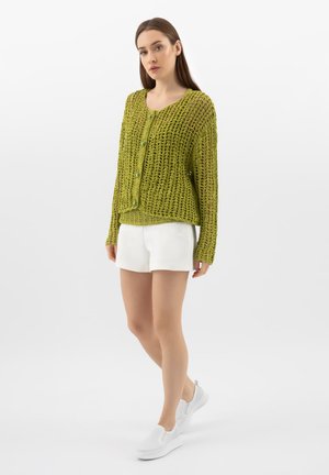 Vrouw die een losse, opengebreide groene cardigan met groene knopen draagt, witte shorts en witte instapsneakers, staand tegen een effen witte achtergrond.