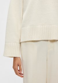 Krém színű pulóver bordázott szegéllyel és mandzsettával, puha anyagból készült. Világos színű, sima tapintású nadrággal párosítva.