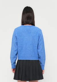 Blauer gestrickter Pullover mit rundem Halsauschnitt und langen Ärmeln, kurz geschnitten, kombiniert mit einem gefalteten schwarzen Rock. Weiche Textur sichtbar.