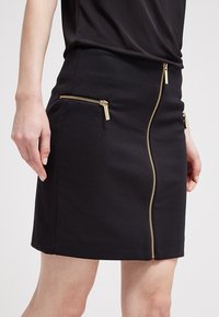 Jupe mini noire en tissu extensible, dotée d'une fermeture éclair dorée diagonale et de deux poches zippées à l'avant. Texture lisse.
