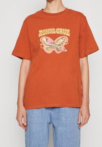 Rost-orange bomullströja med en grafisk fjärilsdesign i rosa och gult, med stjärnor och texten "ANTA CRUZ" i en retrostil.