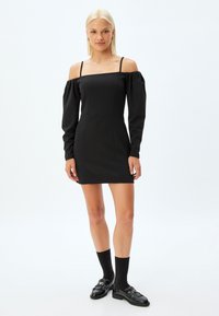 Schwarzes schulterfreies Minikleid mit langen Puffärmeln, tailliertem Oberteil und glatter Textur. Ergänzt mit schwarzen Loafers und knielangen Socken.