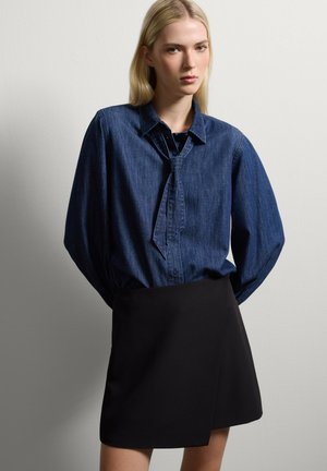Camicia di denim con colletto strutturato e dettaglio cravatta, abbinata a una gonna avvolgente nera, su uno sfondo grigio chiaro.