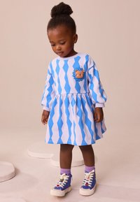 Haljina plave i bijele cik-cak uzorka s dugim rukavima, s aplikacijom lava na prsima, uparena s plavim tenisicama i ljubičastim čarapama.