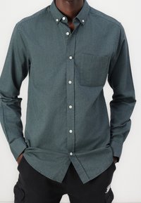 Uomo con una camicia verde scuro a bottoni con tasca sul petto e pantaloni neri, mani nelle tasche, su uno sfondo bianco semplice.