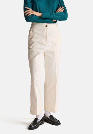 Cremfarbene Hosen mit hoher Taille, vorderer Knopfschluss und Seitentaschen; kombiniert mit schwarzen Loafern und weißen Socken.