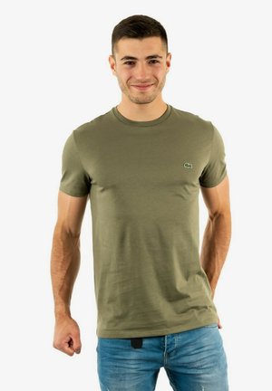 T-shirt in cotone verde oliva con maniche corte, scollatura rotonda e un piccolo logo sul lato sinistro del petto, abbinato a jeans blu.