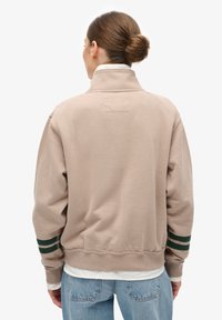 Sweatshirt beige avec un col haut, des poignets côtelés et des accents rayés verts sur les manches. Design simple et texture douce, adapté à un usage décontracté.