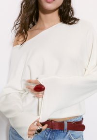 Off-white asymetrický sveter s dlhými rukávmi, nosený s modrými džínsami s vysokým pásom a červeným textúrovaným opaskom. Obsahuje veľký červený prsteň.