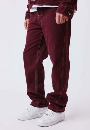 Gambe con pantaloni larghi color bordeaux con cuciture bianche e scarpe da ginnastica bianche, in piedi davanti a uno sfondo semplice.