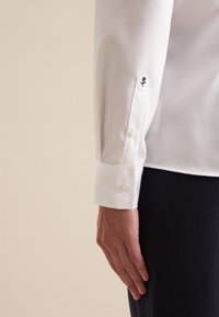Camicia bianca a maniche lunghe con polsini abbottonati e un piccolo ricamo di rosa nera vicino al polsino, abbinata a un tessuto scuro sotto.