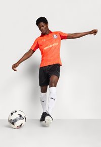 Oranžna športna majica z rdečimi detajli, črne kratke hlače, bele nogavice in črne kopačke. Igralec je v gibanju, s bližnjim nogometnim žogom.