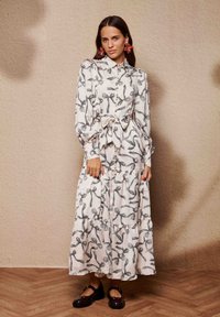 Φόρεμα με μακριά μανίκια και κουμπιά σε κρεμ χρώμα με σχέδιο από μαύρες κορδέλες. Διαθέτει ζωνάρι στη μέση, φούστα με στρώσεις και συνδυάζεται με σκουρόχρωμα παπούτσια.