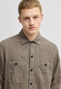 Lichtbruine linnen button-up shirt met een puntige kraag, zwarte knopen en twee borstzakken. Zachte textuur met een relaxte pasvorm.