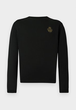 Črna majica z dolgimi rokavi iz mehkega blaga, s majhnim zlatim vezenim grbom na levi strani prsnega koša, okroglim izrezom in oprijetimi rokavi.