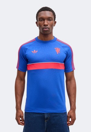 Άνδρας που φοράει μπλε μπλουζάκι Adidas με κόκκινες ρίγες στα μανίκια, κόκκινη οριζόντια ρίγα στο στήθος και λογότυπα στο πάνω μέρος του στήθους.