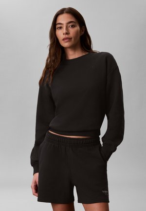 Femeie cu păr lung și brunet, purtând un hanorac negru cu mânecă lungă și pantaloni scurți negri asortați, stând pe un fundal gri simplu.