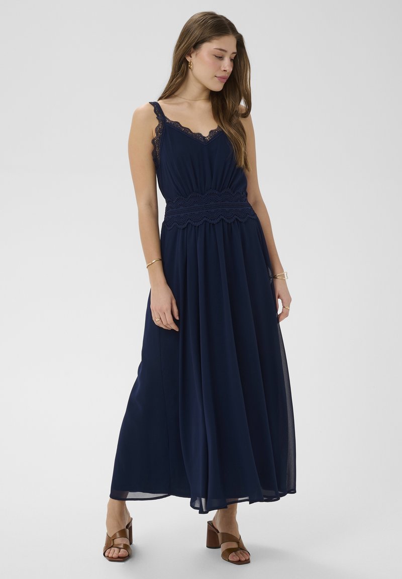 Junge Frau trägt ein ärmelloses, marineblaues Maxikleid mit Spitzendetails, steht und schaut nach unten, dazu braune Sandalen mit Absatz.