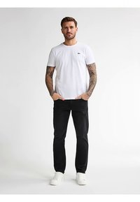 Bílé bavlněné tričko s krátkými rukávy a malým logem na hrudi, doplněné černými džínami a bílými teniskami. Na pažích jsou viditelné tetování.