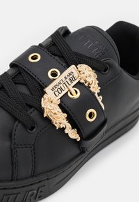 Baskets noires avec lacets noirs, œillets dorés et boucle décorative dorée Versace Jeans Couture sur la bride avant.