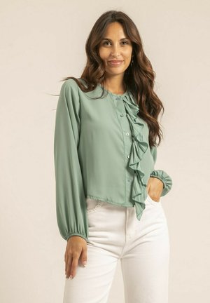 Blouse verte et légère avec un devant à volants, fermeture à boutons et manches longues bouffantes, assortie à un pantalon blanc taille haute.