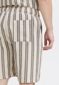 Beige shorts with black vertical stripes, made of textured fabric. Features a back pocket on the right side and an elastic waistband.