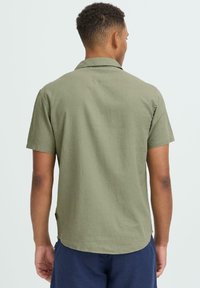 Chemise à manches courtes de couleur vert olive avec une coupe décontractée et un tissu subtilement texturé. Elle présente un col classique et un ourlet arrondi.