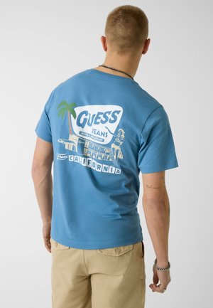 Hombre con camiseta azul de Guess Jeans con gráfico de un motel de California, palmera y bailarina de hula, pantalones beige y pulsera de cadena plateada.