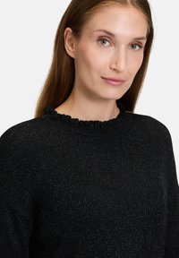 Sort tekstureret sweater med en ribbet design og en dekorativ volangæde. Har subtile metalliske tråde for ekstra visuel detalje.