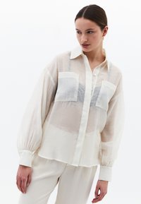 Lekka, zwiewna koszula w kremowym kolorze z zapięciem na guziki z przodu, długimi rękawami oraz dwiema kieszeniami na piersi z szarymi akcentami. Gładka faktura na całej powierzchni.