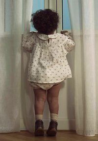 Šviesios spalvos, tekstūruoto audinio marškiniai su apvaliu apykakle ir mažais raštais. Dera su atitinkančiomis kelnaitėmis ir tamsiais batais. Vaikas stovi šalia lango.