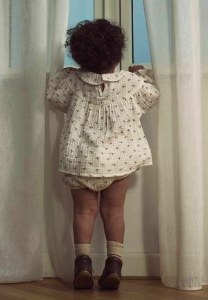 Ljusfärgad, texturerad skjorta med rund krage och små mönster. Sammanställd med matchande byxor och mörka skor. Barnet står vid ett fönster.